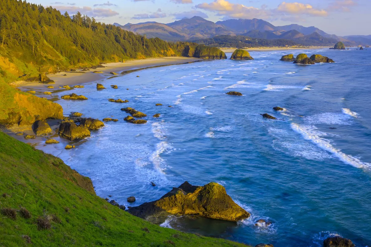 A sweeping view of the rocky coastline with waves crashing against the shore, distant mountains, and a sandy beach in the foreground. The vista captures the natural beauty and tranquility of the Oregon coast, inviting exploration and outdoor enjoyment.