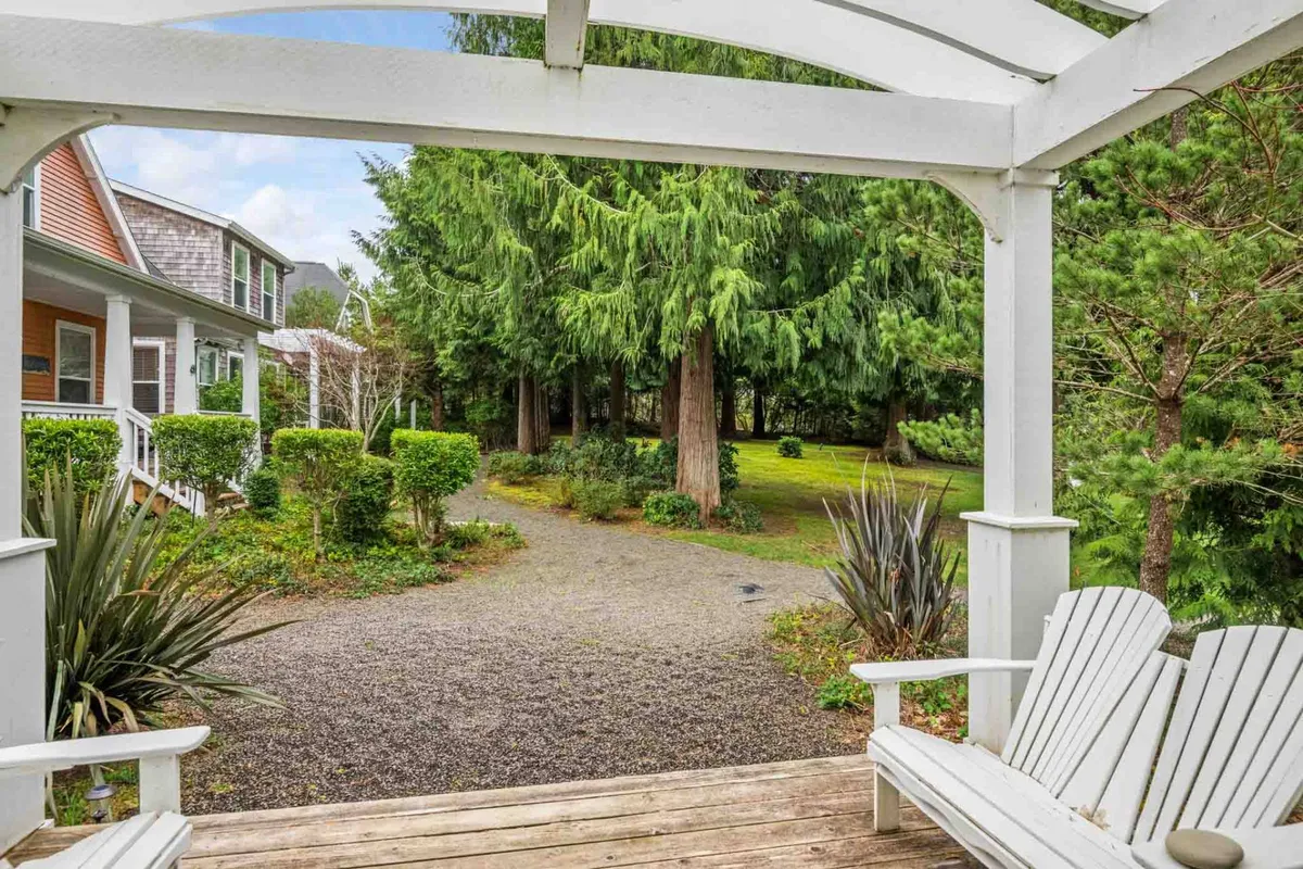 A tranquil exterior scene with white wooden chairs set along a gravel pathway, enveloped by lush greenery. The inviting porch suggests a space perfect for morning coffee or evening conversations, while the garden setting evokes a peaceful, social atmosphere ideal for friends gathering outdoors.