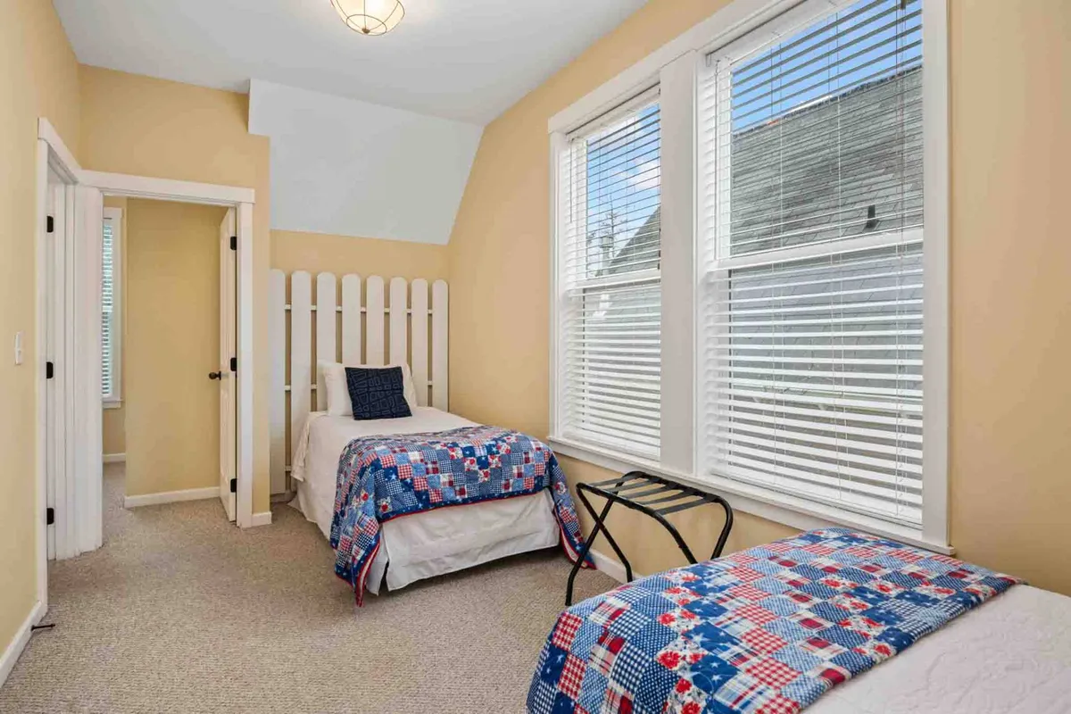 A bright and welcoming sleeping nook with two single beds adorned with colorful quilts, positioned beside large windows that let morning light stream in. A decorative white wooden partition subtly separates the space, making it cozy yet connected.