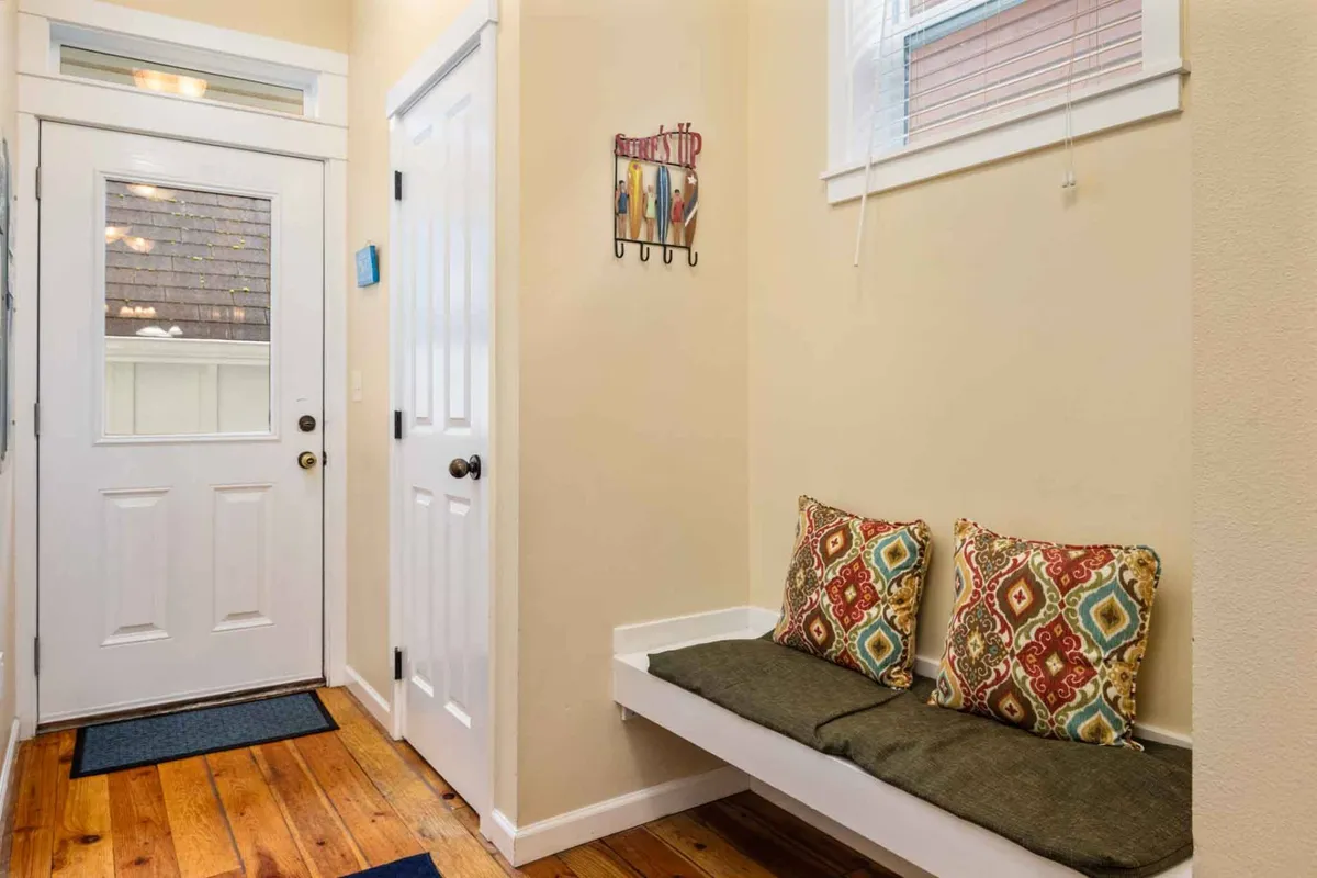 A bright and welcoming entryway with a cushioned bench, decorative wall art, and the front door in view. The area is organized and inviting, offering a first impression of warmth and hospitality.