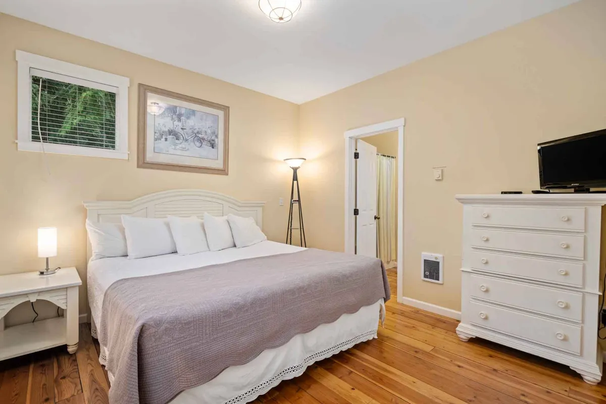 A warm and inviting bedroom featuring a comfortable bed, dressing table, lamp, and artwork. Sunlight streams through the windows, creating a restful atmosphere ideal for relaxation or quiet moments. The space offers both privacy and accessibility, making it suitable for adults seeking a retreat.