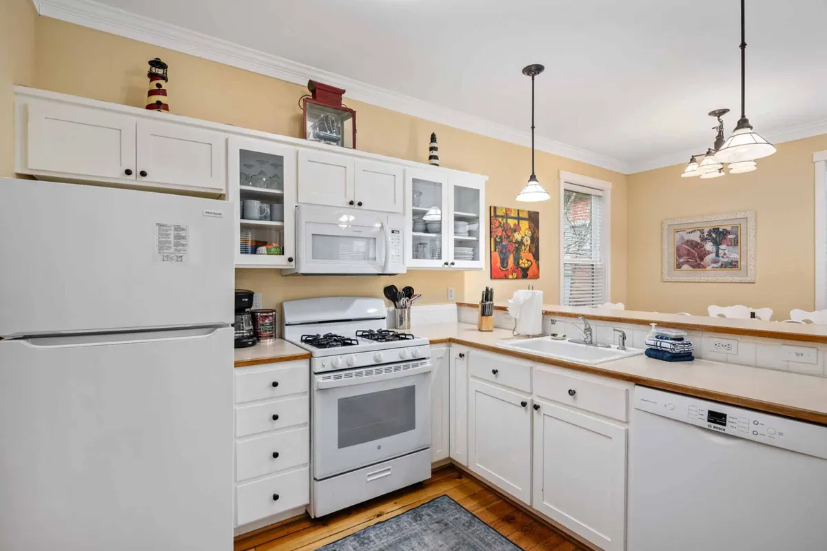 The image highlights a kitchen with white cabinetry, warm wooden countertops, colorful artwork, and a dining area visible in the background—evoking a sense of warmth and togetherness. The setup is designed for both cooking and entertaining.
