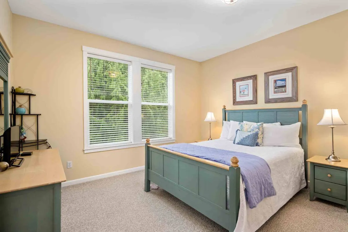 A serene bedroom with a bed, nightstand, dresser, and large windows letting in ample daylight. The neutral palette and simple decor create a calming ambiance, perfect for a restful night's sleep or quiet family time.