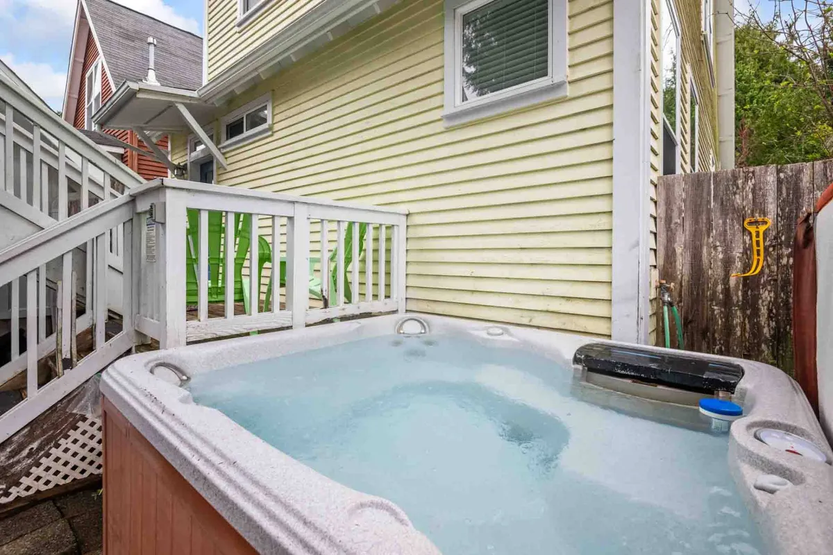 A bubbling hot tub sits on the deck, framed by green chairs and the house’s cheerful yellow siding. A staircase leads up to the area, and the setting is tranquil, perfect for unwinding while enjoying the outdoors.