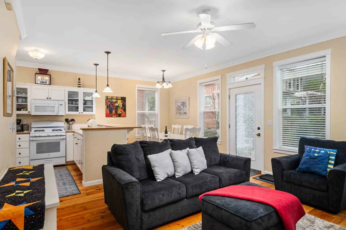 This open-plan interior image showcases a cozy sofa with decorative pillows, a spacious dining area, visible kitchen appliances, large windows allowing in ample natural light, and a textile accent on the bench. The space appears welcoming and ideal for both casual and festive gatherings.