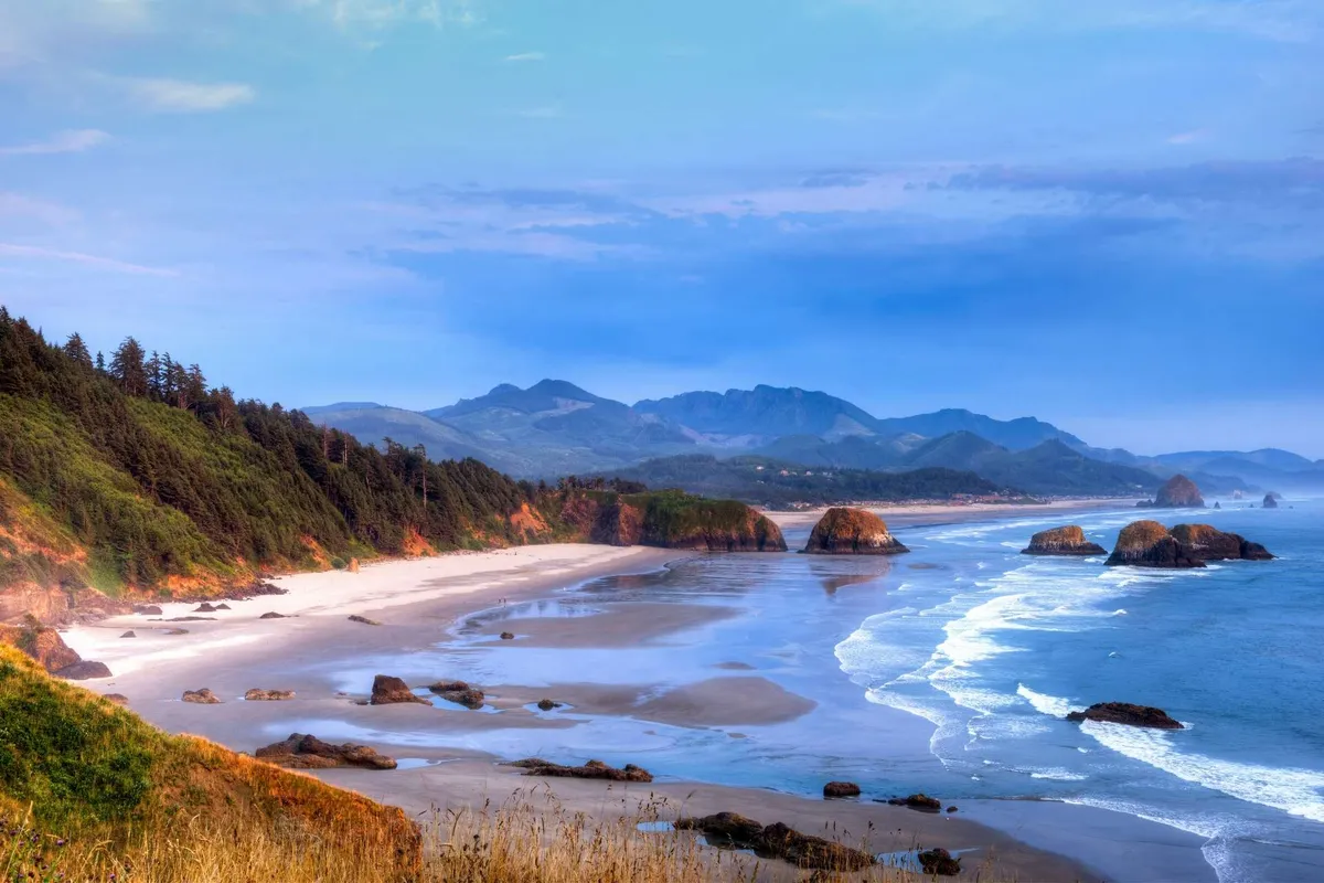 A sweeping coastal view featuring ocean waves rolling onto a sandy beach, framed by rock formations and distant mountains. The tranquil atmosphere and expansive shoreline capture the allure of a sunset stroll and the area's natural beauty.