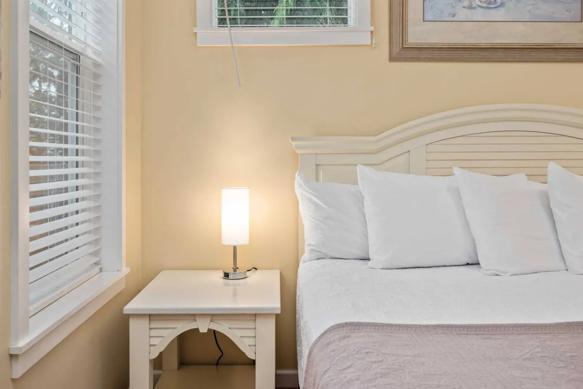 A peaceful bedroom with a cozy bed dressed in white linens, a nightstand with a lamp, and framed artwork. Sunlight filters through a window, creating a serene atmosphere ideal for relaxation and rejuvenation.
