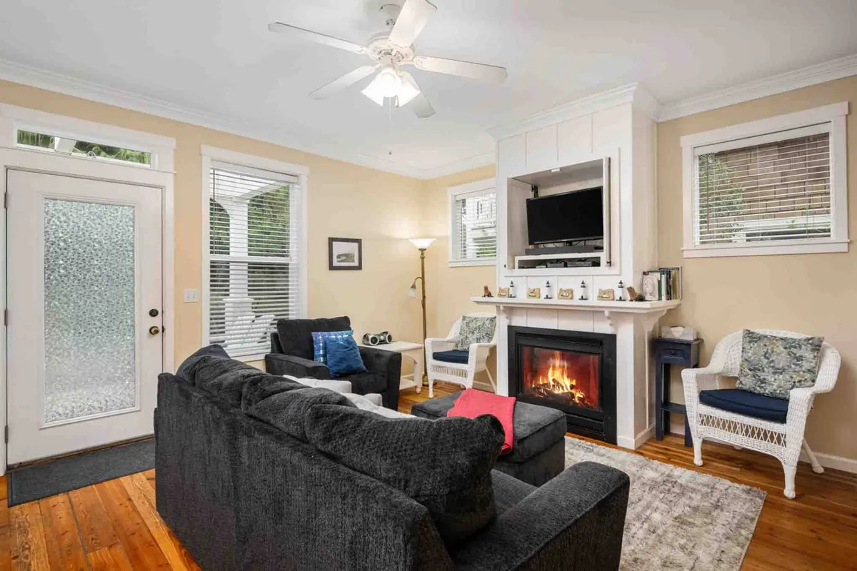 This living area features a prominent fireplace, a comfortable sofa, large windows allowing in sunlight, and a cozy lamp. The inviting setting is perfect for relaxing, reading, or gathering for conversations and movie nights.